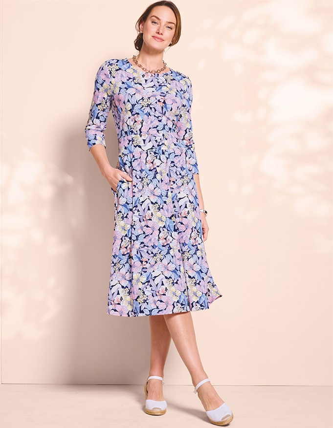 model wearing pruple, blue and white floral dress and white shoes, with one hand in a pocket