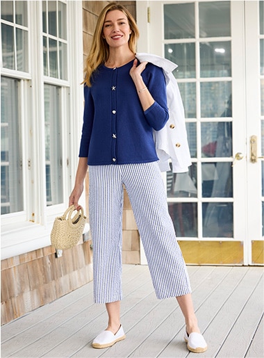 model wearing teal jacket and top over white pants and shoes holding a tan hat with white rim